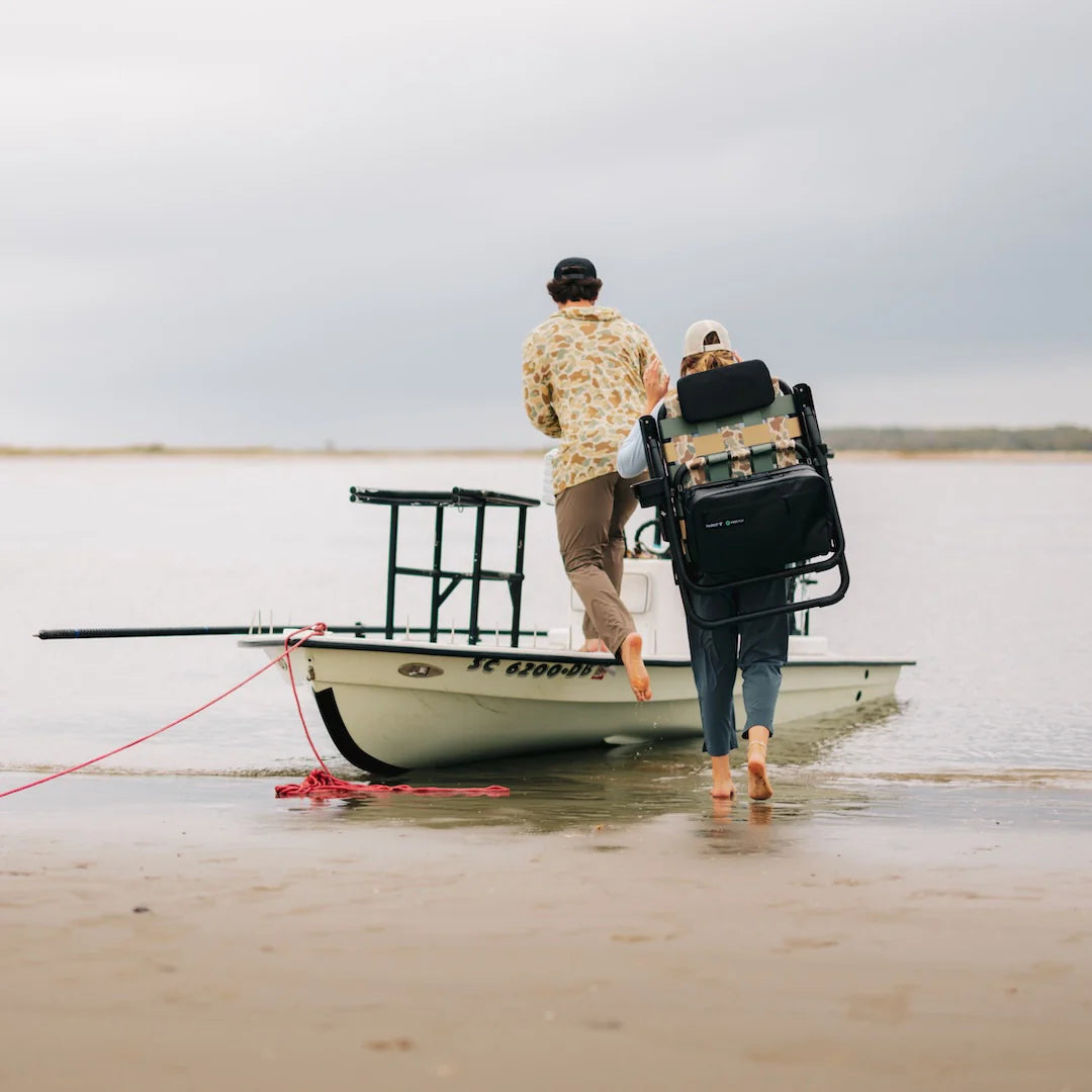 Two people walking barefoot toward a skiff, one carrying the PARKIT Free Fly Eclipse Recliner in Vintage Camo using its backpack carry straps. The folding outdoor chair features an integrated cooler and rugged aluminum frame the ultimate portable beach, boat, and camping chair for couples, adventurers, and weekend explorers.